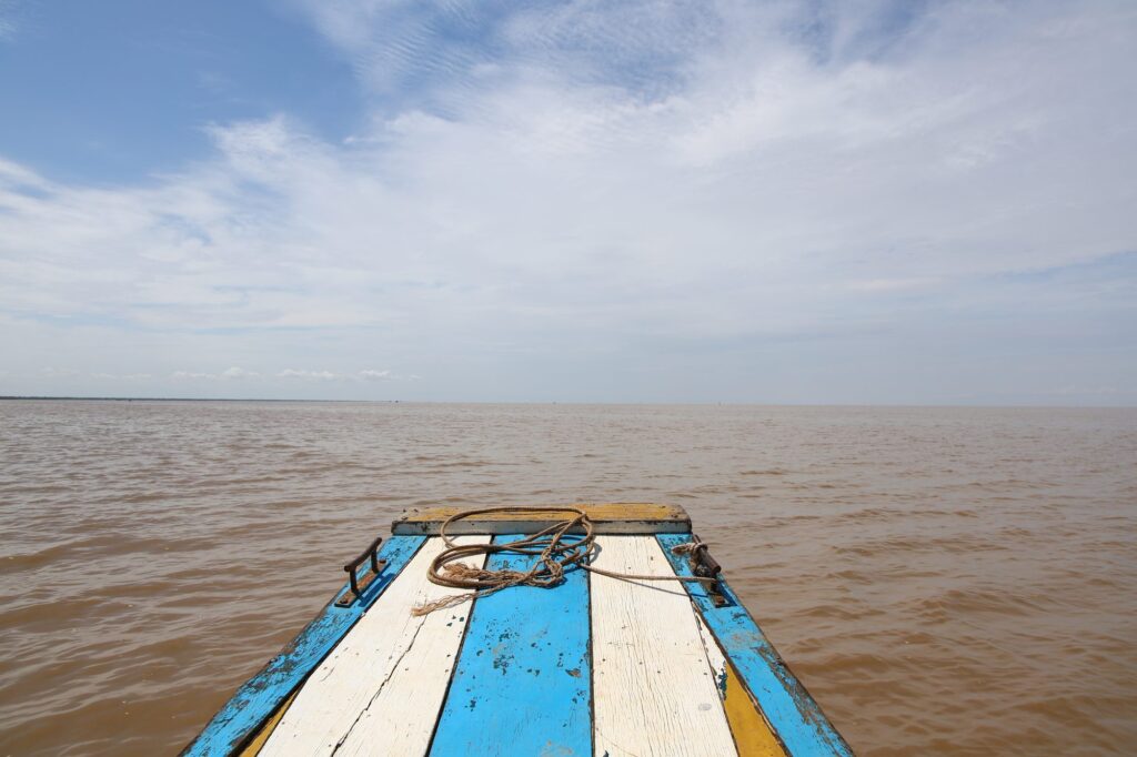 The bow of a blue and white wooden boat heading out onto the brown water of Tonle Sap lake